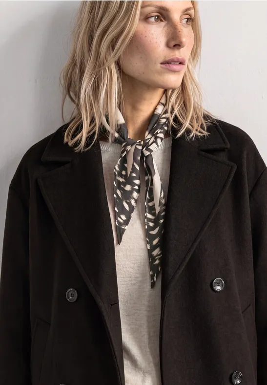 Eine blonde Frau in schwarzem Mantel, grauem Pullover und gemustertem Schal blickt seitlich.