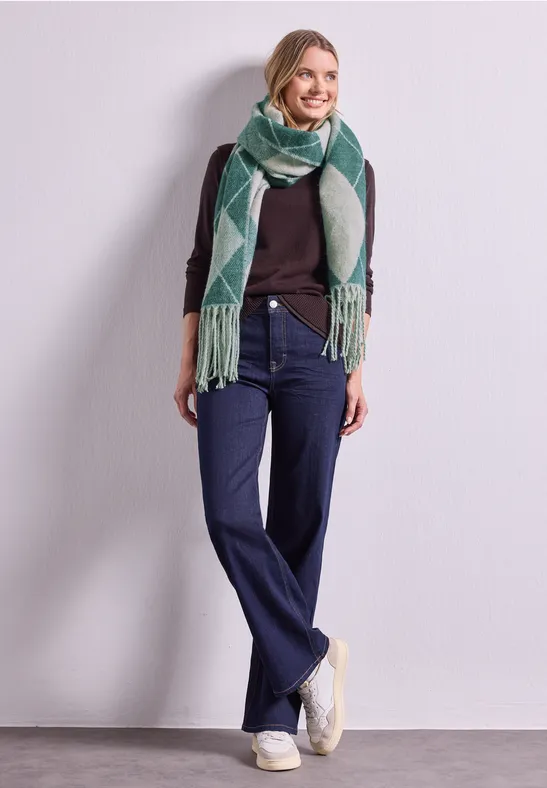Frau in lässigem Outfit: dunkler Pullover, weite Jeans, weiße Sneaker und grüner Schal mit Muster vor neutralem Hintergrund.