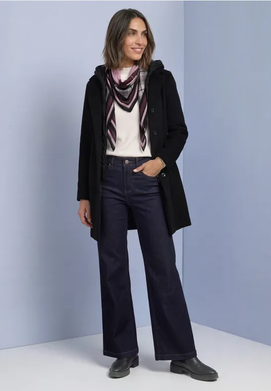 Frau in schwarzer Jacke, helles Oberteil, Schal und weiten Jeans, steht lächelnd vor hellem Hintergrund.