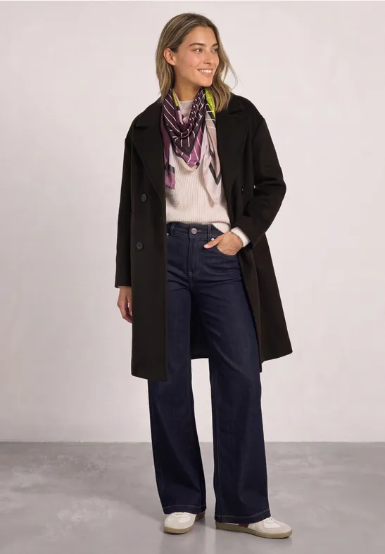 Frau in schwarzem Mantel, weißem Pullover, weiten Jeans, weißen Schuhen und gemustertem Schal.