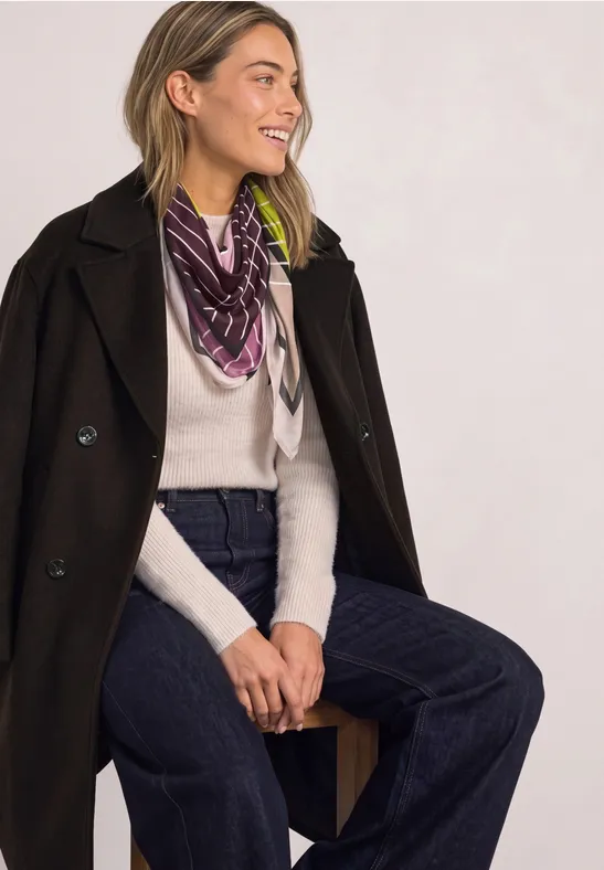 Eine lächelnde Frau sitzt seitlich, trägt dunklen Mantel, weißen Pullover, Halstuch und weite Jeans.