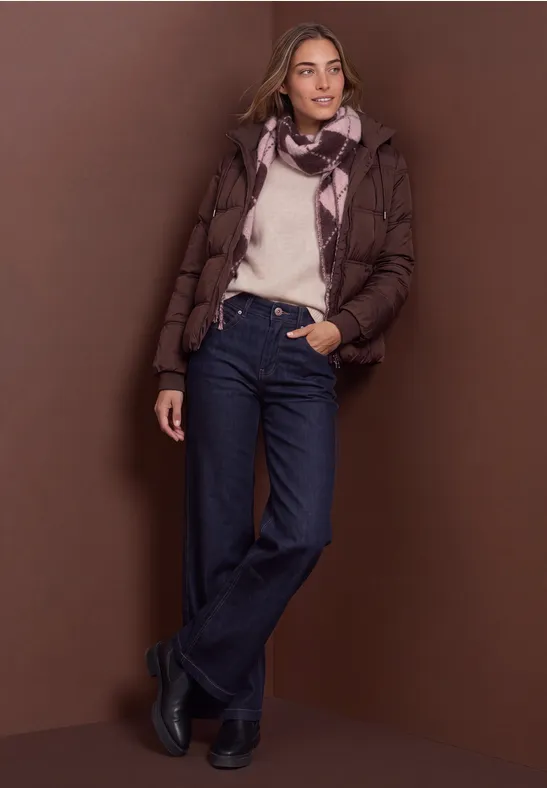 Frau in brauner Steppjacke, weißem Pullover, Jeans und Schal vor braunem Hintergrund.