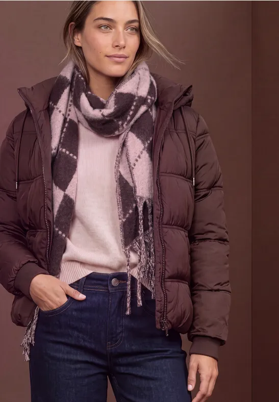 Frau trägt braune Daunenjacke, beigen Pullover, gemusterten Schal und Jeans vor braunem Hintergrund.