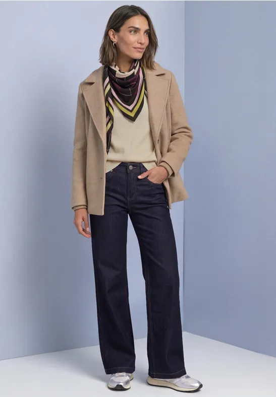 Frau in beiger Jacke, cremeweißem Pullover, farbigem Tuch, weiter Jeans und weißen Sneakern.