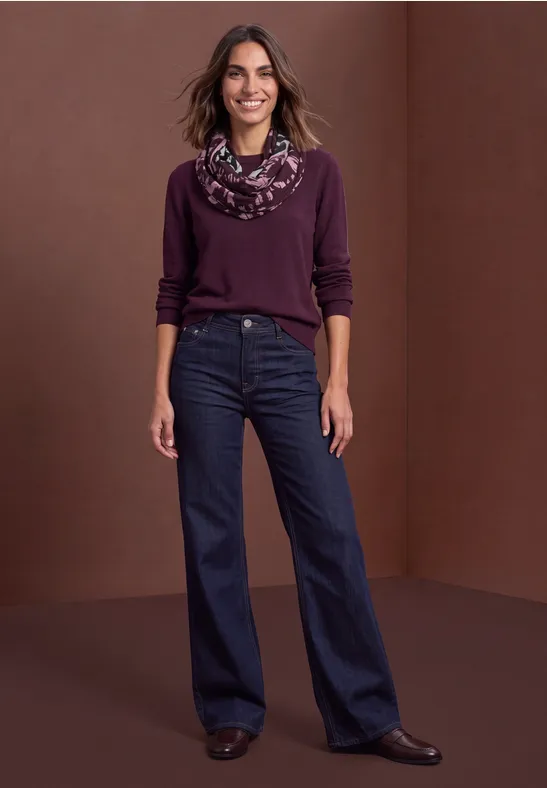 Eine Frau in lila Pullover, gemustertem Schal und dunklen Jeans steht vor braunem Hintergrund.