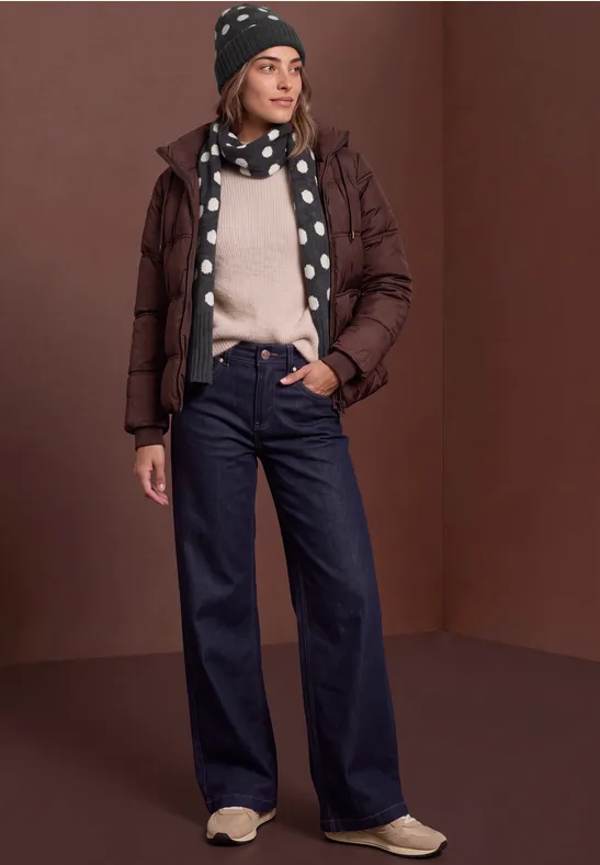 Frau in brauner Daunenjacke, gepunktetem Schal & Mütze, hellem Pullover, weiten Jeans und Sneakers.