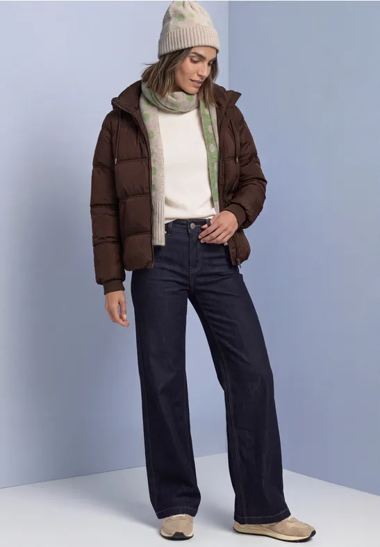 Frau in brauner Steppjacke, weißem Pullover, weiter Jeans und Mütze steht vor hellblauem Hintergrund.