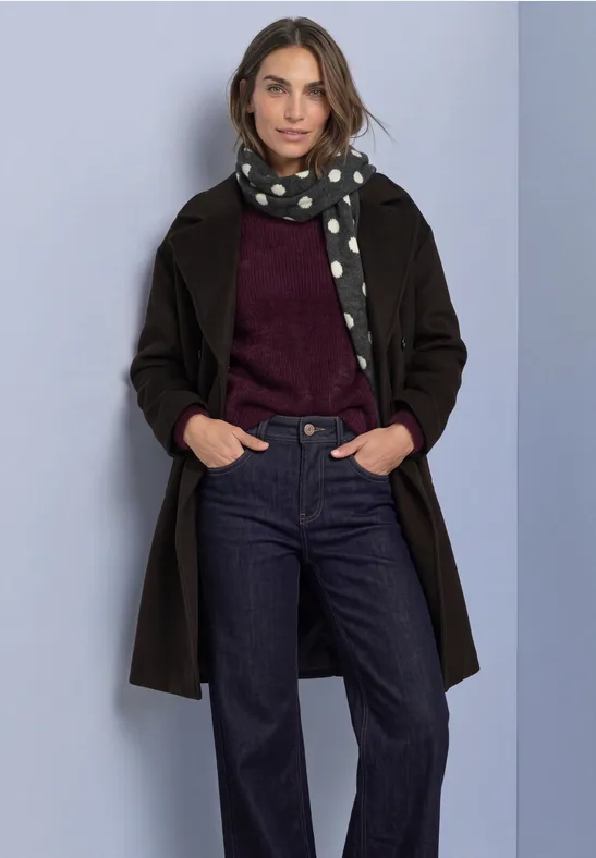 Vrouw in zwarte mantel, gestippelde sjaal, bordeaux trui en donkere jeans, handen in zakken, lichte achtergrond.
