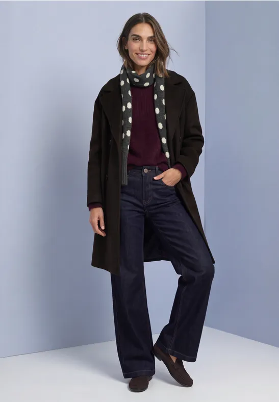 Een lachende vrouw in een lange jas, bordeaux trui, wijde jeans en een stippen-sjaal voor een neutrale achtergrond.