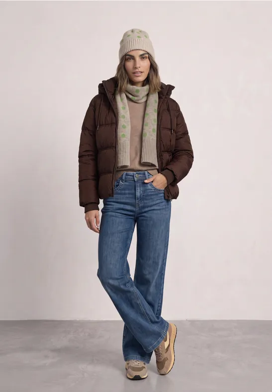 Frau in brauner Steppjacke, Jeans, beiger Mütze und Schal, steht vor hellem Hintergrund.