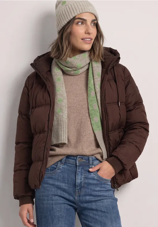 Frau in brauner Daunenjacke, beiger Strickmütze, grün gemustertem Schal, beigem Pullover und blauen Jeans.