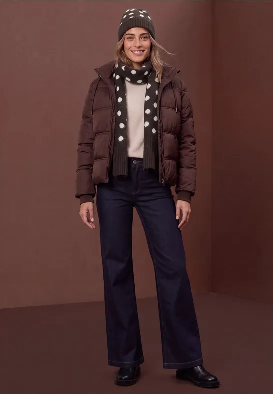 Lächelnde Frau in brauner Daunenjacke, gepunktetem Schal, Mütze und dunklen Jeans vor braunem Hintergrund.