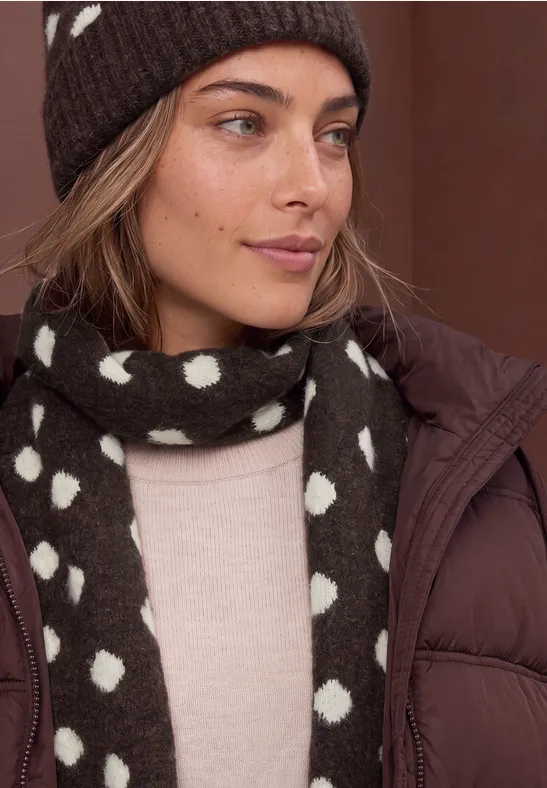 Frau mit brauner Mütze und gepunktetem Schal, beigem Pullover und brauner Daunenjacke.