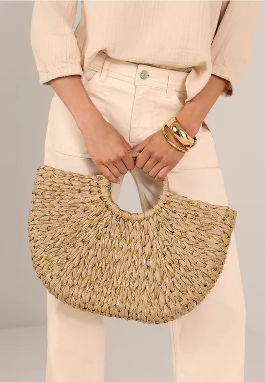 Frau in beige Kleidung hält handgeflochtene Strohtasche in Halbmondform mit Rundgriffen.