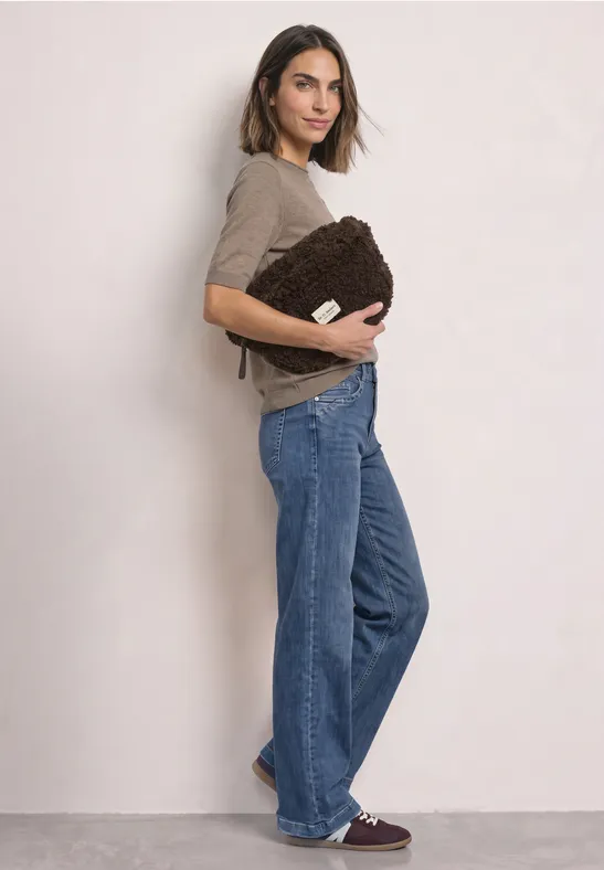 Frau im Profil hält eine braune Plüschtasche, trägt taupefarbenes Oberteil, weite Jeans und dunkelrote Sneaker.