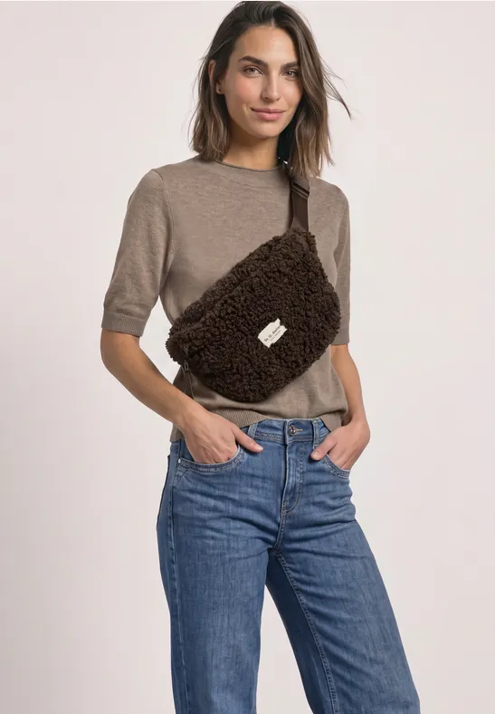 Eine Frau trägt einen beigen Kurzarm-Pullover, blaue Jeans und eine braune, flauschige Bauchtasche vor neutralem Hintergrund.