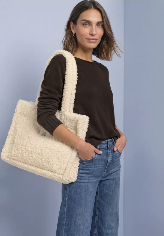 Frau im dunkelbraunen Pullover und Jeans mit flauschiger cremefarbener Tote-Bag vor hellem Hintergrund.