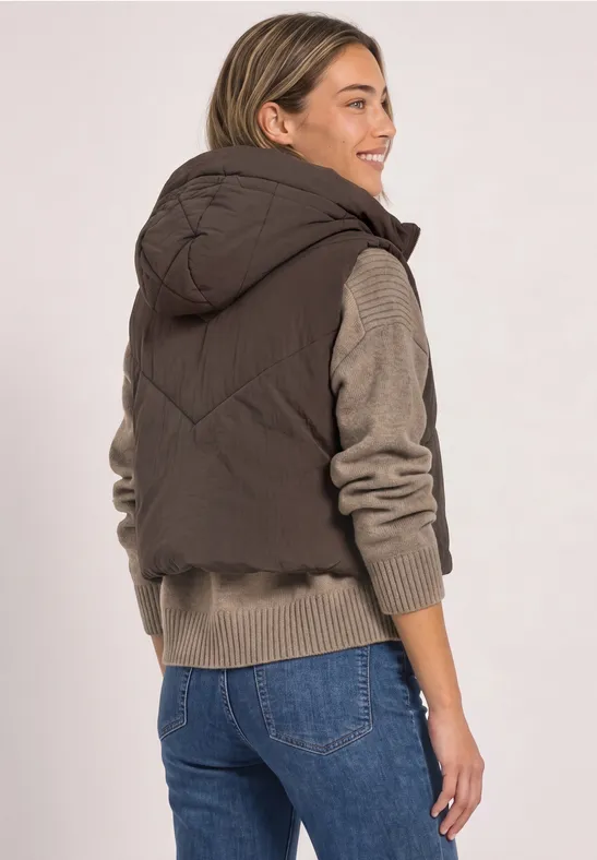 Eine Frau von hinten, die eine braune Steppweste mit Kapuze und Jeans trägt und seitlich lächelt.