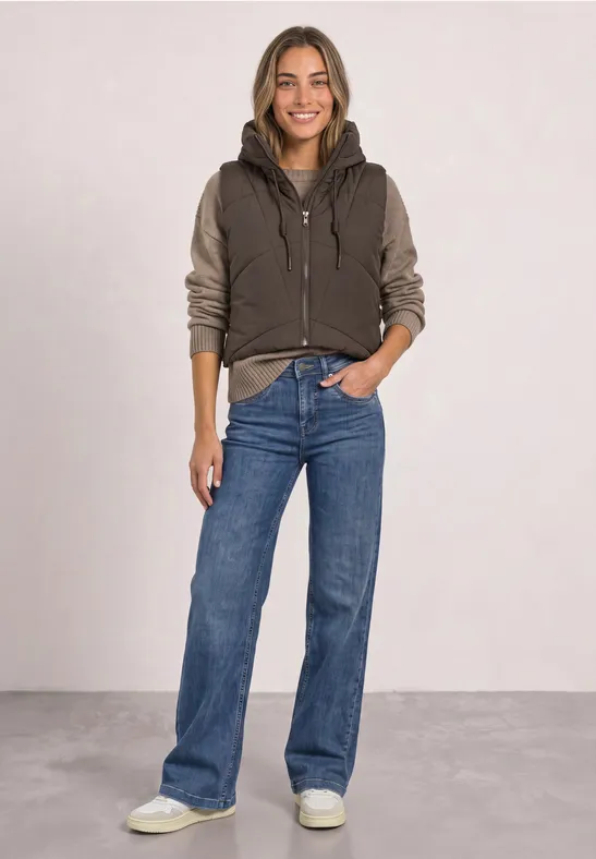 Lächelnde Frau in brauner Jacke mit Hoodie, weiten blauen Jeans und weißen Sneakers, Hände in den Taschen.