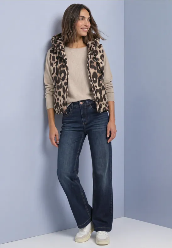 Frau in legerem Outfit: Leo-Weste, helles Shirt, weite Jeans und weiße Sneaker vor grauem Hintergrund.