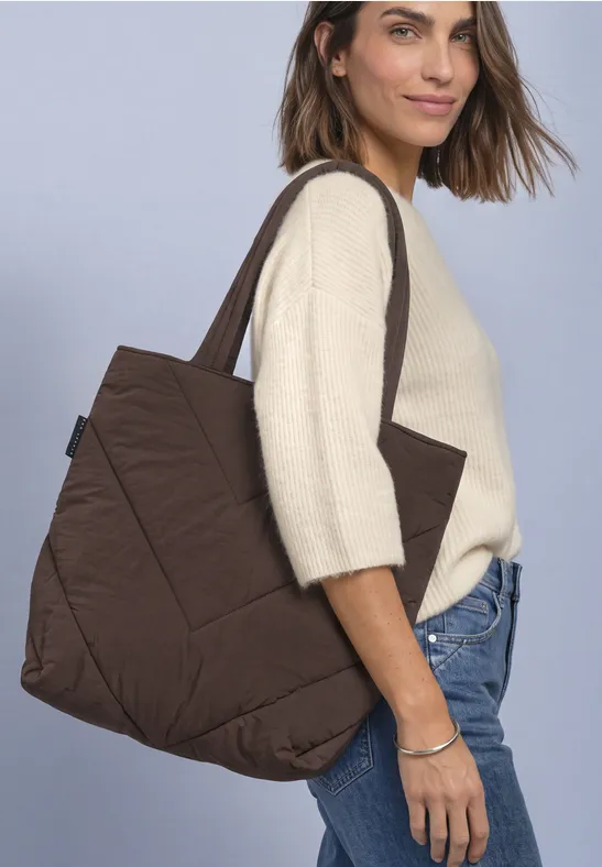 Frau mit beigem Pullover und blauen Jeans trägt große, braune, gesteppte Tote Bag über der Schulter.