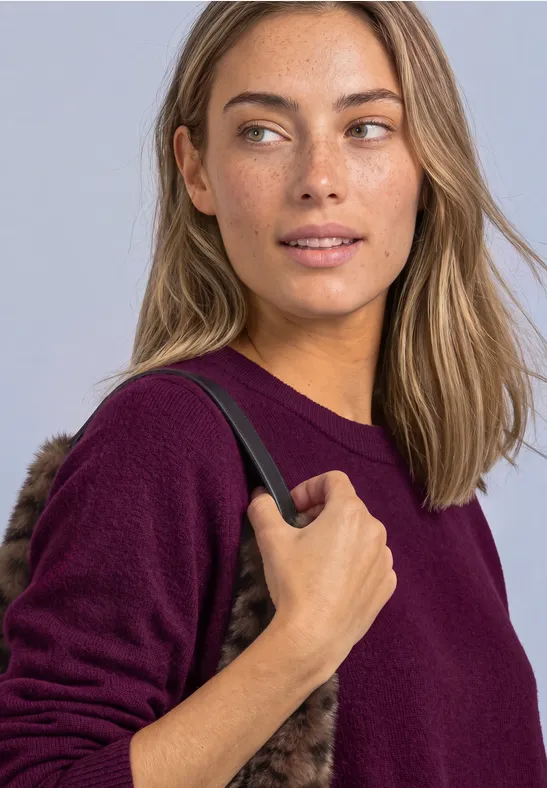 Frau mit Sommersprossen, schulterl. Haaren und lila Pullover hält den Riemen einer schwarzen Tasche.