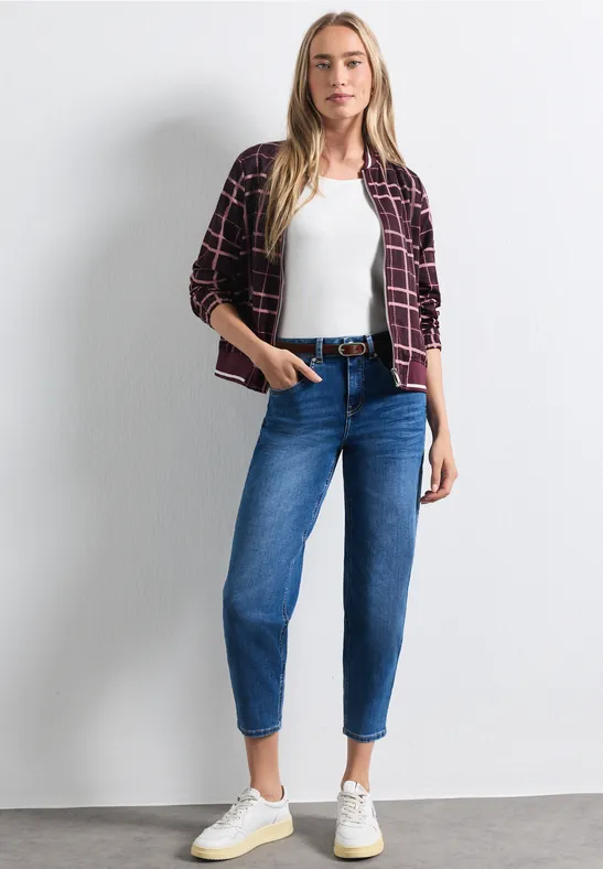 Frau mit kariertem Hemd, weißem Top, blauen Jeans und weißen Sneakers vor hellem Hintergrund