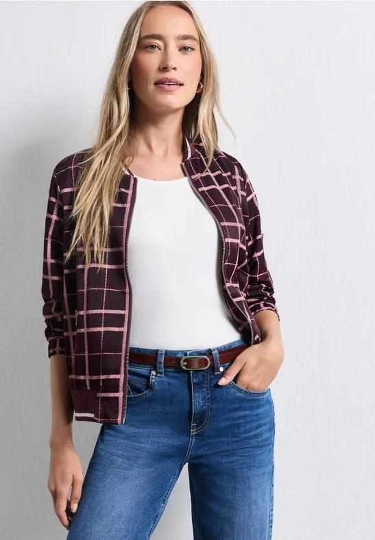 Junge Frau mit blonden Haaren trägt weiße Bluse, karierte Jacke und blaue Jeans mit Gürtel.