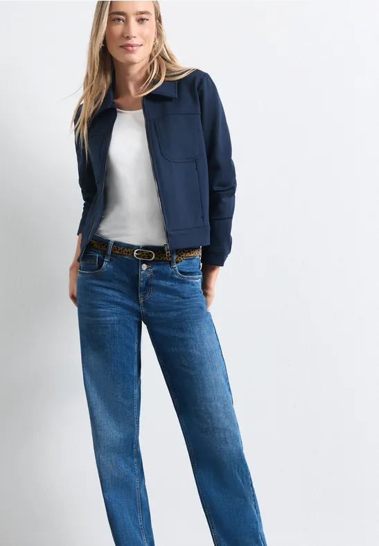 Frau in blauem Blouson, weißem Top und Jeans mit Gürtel vor weißem Hintergrund.