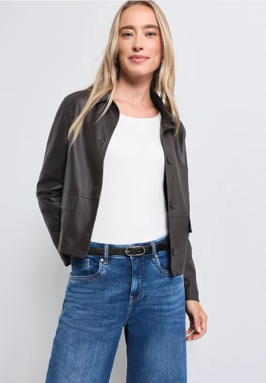 Eine Frau in weißem Top, schwarzer Lederjacke und blauen Jeans mit Gürtel lächelt.