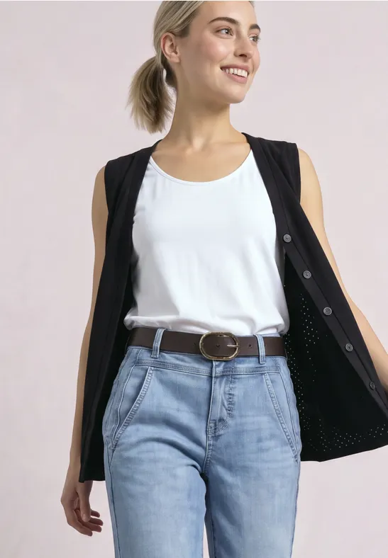 Lächelnde Frau mit blondem Haar, weißem Tanktop, schwarzer Weste offen, hellblauen Jeans und Gürtel.
