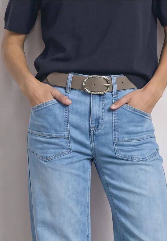 Person mit blauer Jeans, breitem Gürtel und dunklem Oberteil, Hände in den Taschen