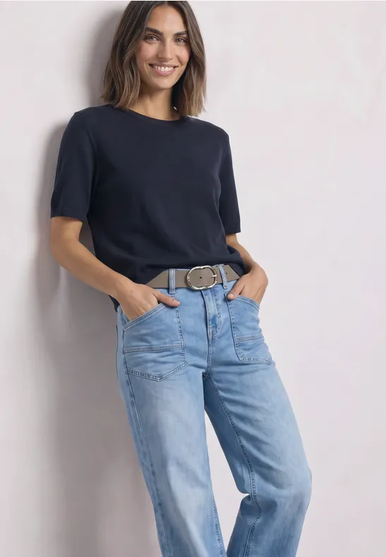 Ein lächelndes Modell in schwarzem T-Shirt und blauen Jeans mit Gürtel.