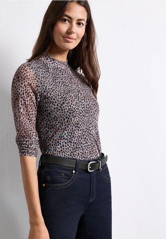 Frau in semi-transparentem Leopardenmuster-Shirt und dunkler Jeans vor weißem Hintergrund.