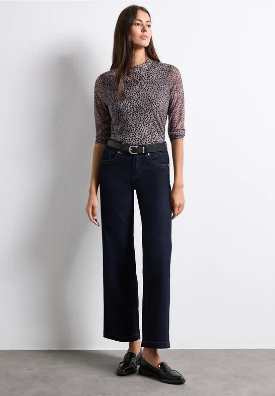 Frau in gemustertem Oberteil und weiten, dunklen Cropped-Hosen, schwarze Loafer.