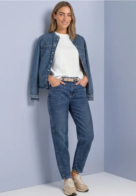 Frau trägt lässiges Jeans-Outfit mit weißem Shirt und Sneakern, steht vor hellvioletter Wand und lächelt.
