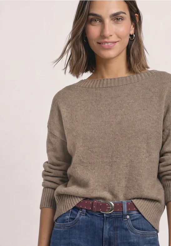 Frau in braunem Strickpullover, blauen Jeans und rotem Flechtgürtel, lächelnd.