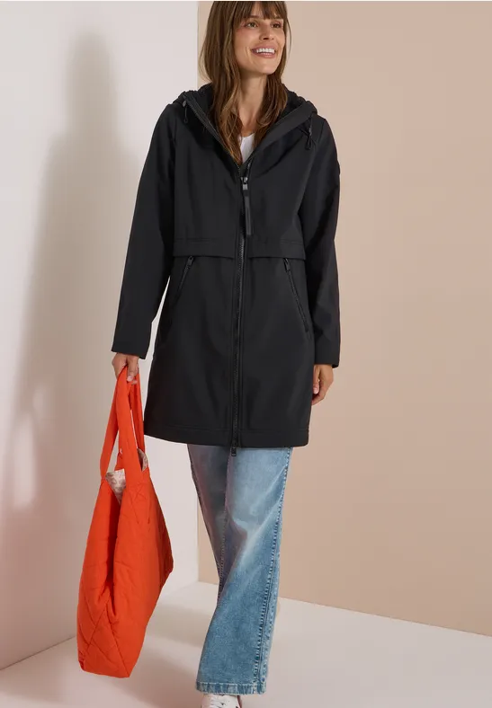 Femme en imperméable noir et jean, souriante, avec un grand sac orange devant un fond uni.