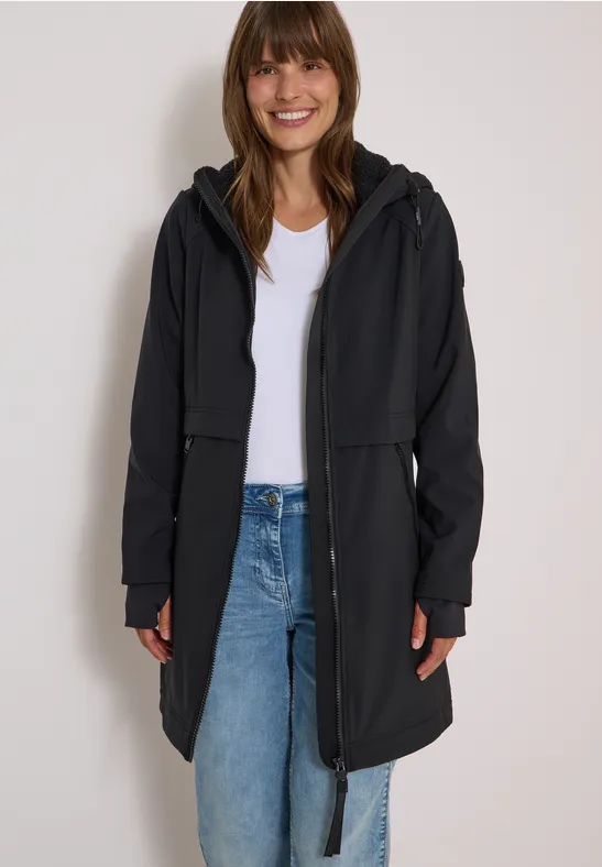 Femme aux cheveux bruns porte un pull blanc, un jean bleu et un long manteau noir, souriante devant un fond clair.