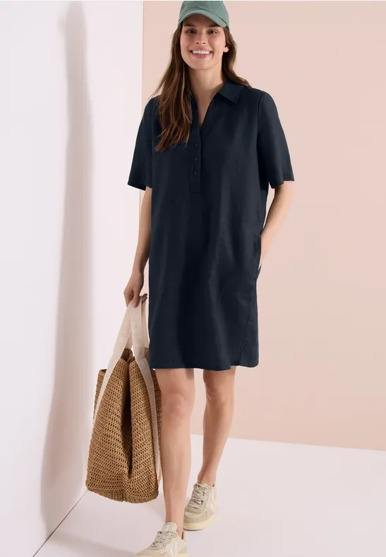 Frau in dunkelblauem Hemdblusenkleid mit Kurzarm, Sneakers und großer Strohtasche.