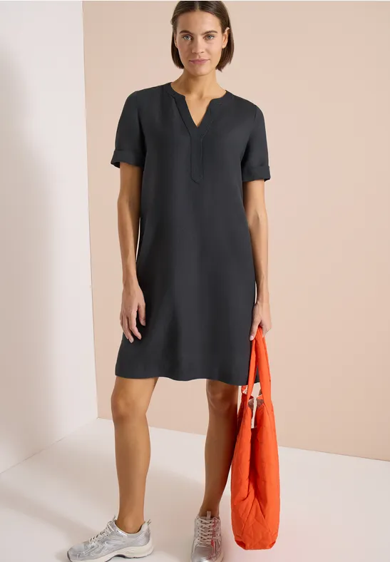 Frau in knielangem, kurzem schwarzen Kleid, weiße Sneaker, orangene Tasche, vor neutralem Hintergrund.