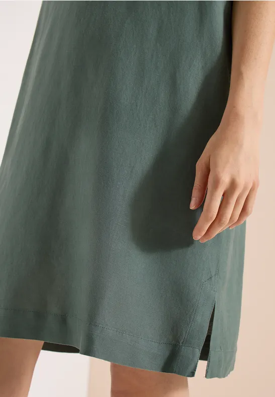 Nahaufnahme: Person trägt grünes, leichtes Leinenkleid mit seitlichem Schlitz, Hand entspannt am Körper.