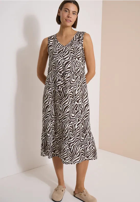 Frau trägt ärmelloses Midikleid mit Zebra-Muster und beigen Loafers vor schlichtem Hintergrund.