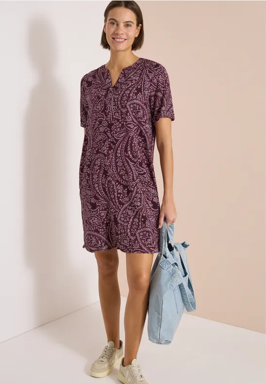 Frau trägt ein kurzes, lila Paisley-Muster-Kleid, weiße Sneaker und hält eine Jeansjacke.