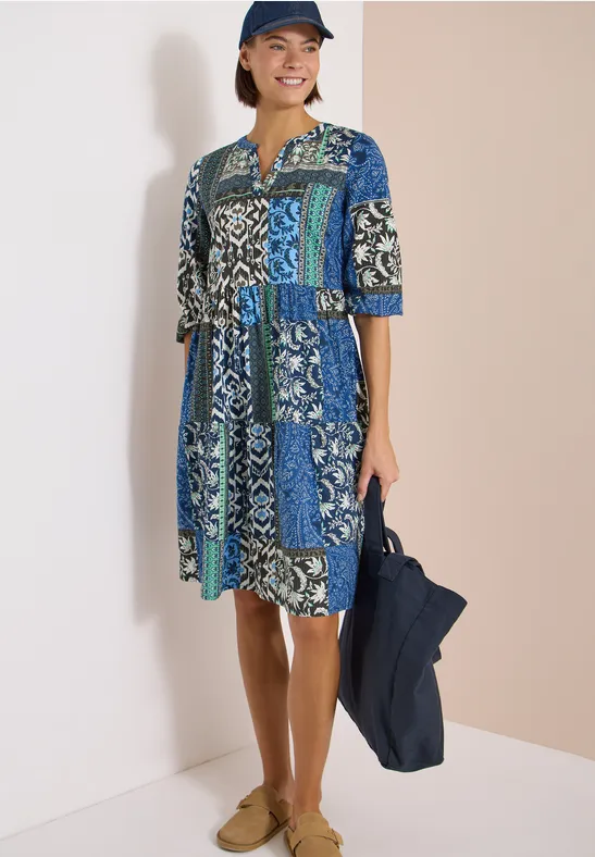 Frau in knielangem, bunt gemustertem Kleid in Blau- und Grüntönen mit navy Tasche und Hut.