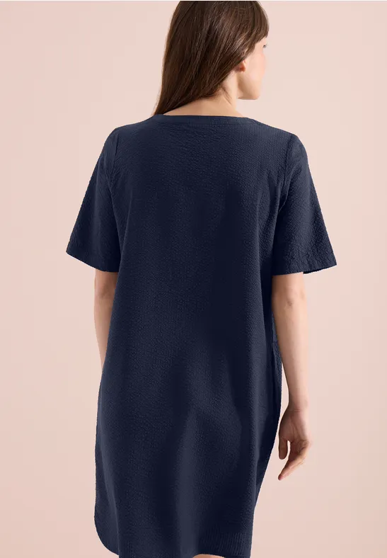 Eine Frau von hinten in einem locker sitzenden dunkelblauen T-Shirt-Kleid vor hellem Hintergrund.