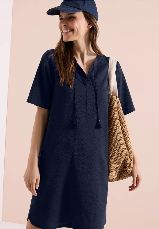 Frau in einem marineblauen, kurzärmeligen Kleid mit V-Ausschnitt und Quasten, mit Cap und Basttasche.