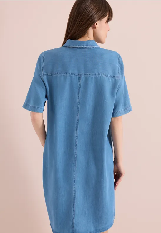 Une femme de dos portant une robe-chemise en denim bleu clair arrivant au genou à manches courtes.