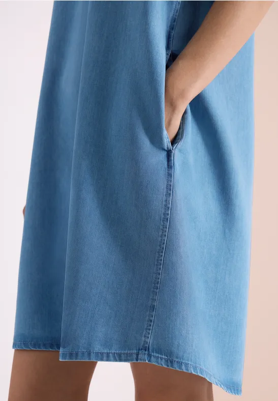 Une personne porte une robe en jean bleu clair, la main dans la poche latérale.