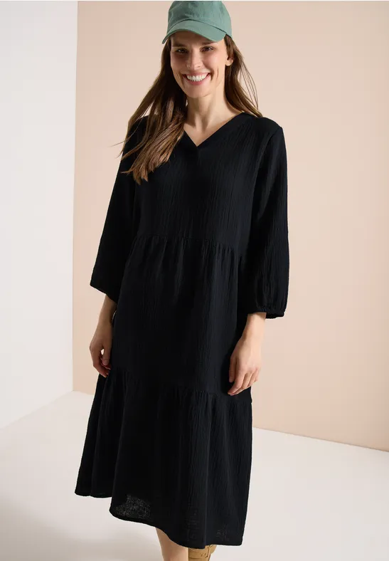 Femme portant une robe midi noire, ample, à encolure en V et manches 3/4, devant un fond clair.